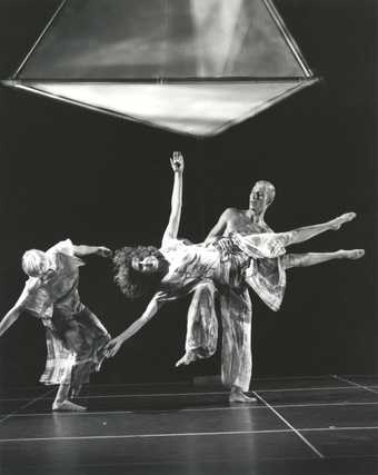 A black and white photo captures three dancers in mid-movement. One dancer is holding another in the air, at hip height, while the third curves around them following the group's movement. All three stand on a stage wearing flowing clothes.