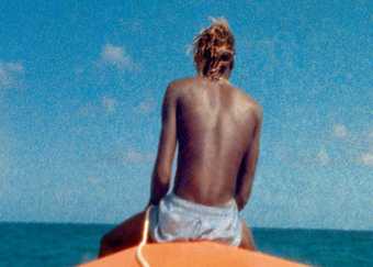 Still from McQueen film showing the back on a man sat on a surf board against a blue sky