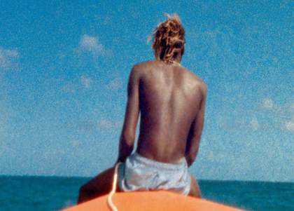 Still from McQueen film showing the back on a man sat on a surf board against a blue sky