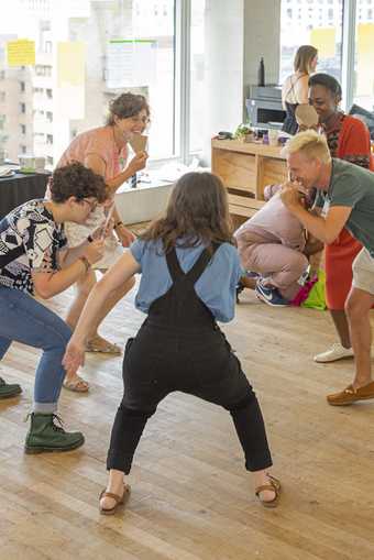 A group of people crouch in a circle facing inwards holding mirrors in front of their faces