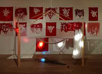 Red and white flags against a white wall; a wooden construction above a wooden branch hung with clothing and beads
