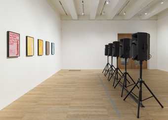 five speakers on stands are arranged in a line facing five paintings hung on a wall. There is a large gap between for a person to walk through.