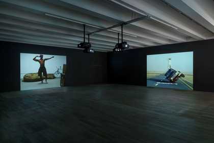 two video projections in a dark room, on the right two people kiss. The left screen is split into four with two pixelated portraits, a blue screen and arabic text