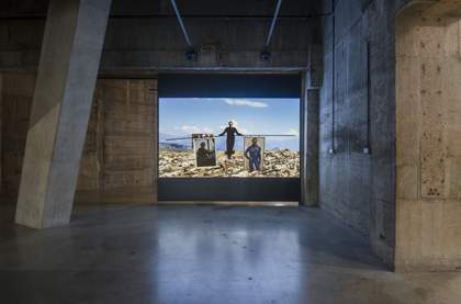 an industrial room with buffed concrete floor and walls. in the centre a film is projected onto a large screen. it reflects on the floor below.