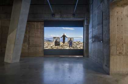 an industrial room with buffed concrete floor and walls. in the centre a film is projected onto a large screen. it reflects on the floor below.