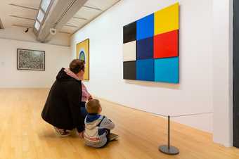 A parent and child crouch on the floor in front of an artwork. There is a wire barrier between them and the artwork.
