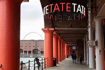 outside of Tate Liverpool on the Albert Dock, the walk way is industrial with big columns and there is a neon sign for Tate overhead. 