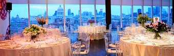 tables laid with white linen and silverware in a large space with windows looking out over the Thames River.