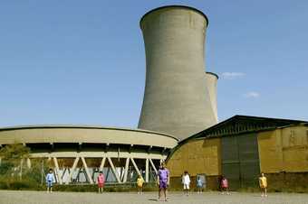 Still from Mikhail Karikis's Children of Unquiet film showing children standing still in front of two large power plant cooling chimneys