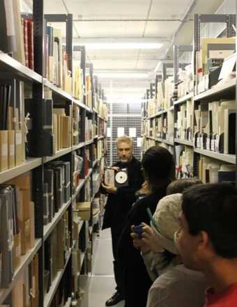 Photograph down a narrow corridor with tall archival shelving on either side; a man speaks to a group, holding up an archival item - a CD or DVD with booklet