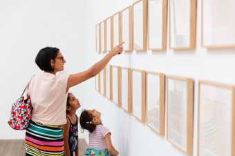 A woman points at art on the wall with her two kids looking on 