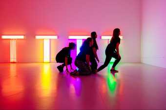 four dancers kneel on the floor and begin to move infront of bright neon lights