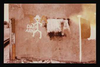 Conrad Atkinson, Colour photograph of a wall in Northern Ireland painted with a stick man, an Irish flag, and the words, ‘Dads Army’ c. 1978