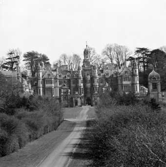 Black and white photograph of huge manor with turrets at the end of a driveway.