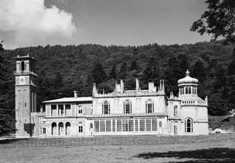 Black and white photograph of a large white manor house with a tower on its left side. This is set against a forest backdrop.