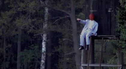 Photograph of a masked performer with one arm outstretched, sitting at the top of a staircase to an elevated shed in a forest