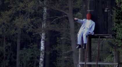 Photograph of a masked performer with one arm outstretched, sitting at the top of a staircase to an elevated shed in a forest