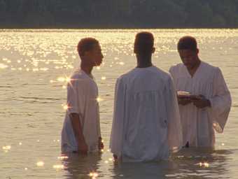A film still with three men standing in the water for a baptism at sunset