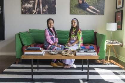 Two girls sat on a sofa wearing traditional dress. the coffee table in front of them has books of Kerry James Marshall and Joan Jonas facing the camera