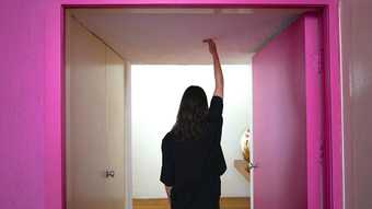 Film still of woman standing in pink a doorway holding an arm to the ceiling
