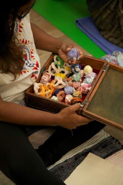 A person sits of the floor with an opened box with thread wrapped around stones inside