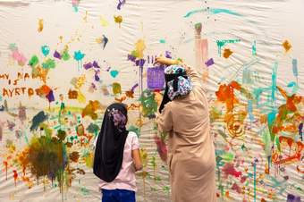 A parent or guardian and child paint on a wall covered in fabric, facing away from the camera