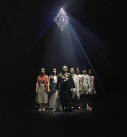 Seven performers stand in a row on a stage, all wearing draped linen clothing in different colours. They are lit by a spotlight appearing out of a large glowing diamond above them.
