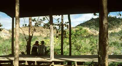 Two boys sit on the wooden floor of a open building on a sunny day