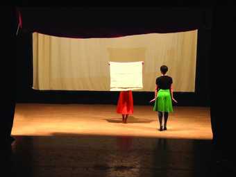 Press Release image of Joëlle Tuerlinckx's work, That's It, featuring two women on stage. One holds white paper over her head