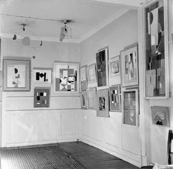 Black and white photograph of a gallery space with geometric paintings hung in frames close together