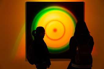 Two silhouetted figures looking at an artwork on the wall of a gallery, glowing orange and green colours in a circular target-shaped form