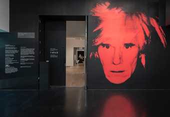 a wall painted black with an open door. to the right of the door is a red print of Andy Warhol. Through the door you can see the exhibition.