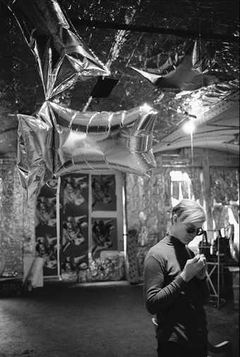 Black and white photograph of Andy Warhol stood below some inflated foil shapes