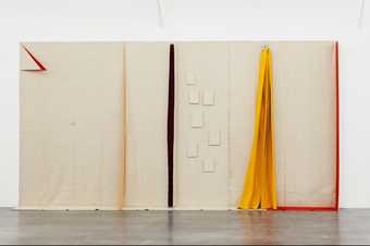 A large canvas hangs from the gallery wall and folds onto the floor. There are slits and cuts which reveal a red colour beneath. On the right side of the canvas hangs a yellow fabric. 