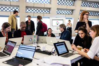 Photograph of a group of people sitting around a table, all looking at laptop screens, in the Tate Exchange space at Tate Modern. In the background, other people stand nearby and watch or talk.