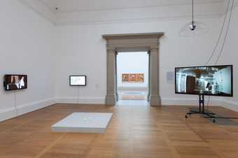 installation shot showing hree TV screens, a floor plinth and a Francis Bacon triptch in the gallery ahead