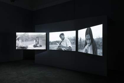 view of three large screens in a dark room, each showing a different black and white image of people in a field