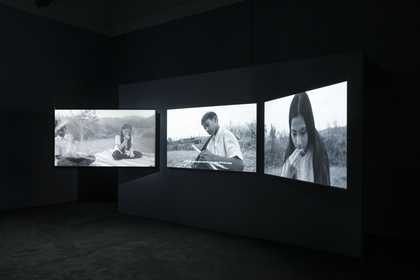 view of three large screens in a dark room, each showing a different black and white image of people in a field