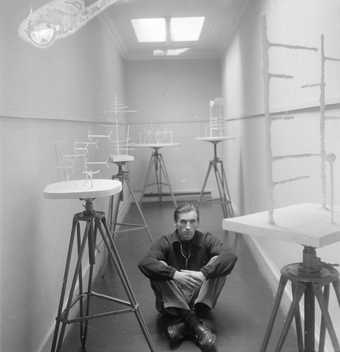 Photograph of William Turnbull in a corridor sitting cross legged on the floor surrounded by wire sculptures on tables