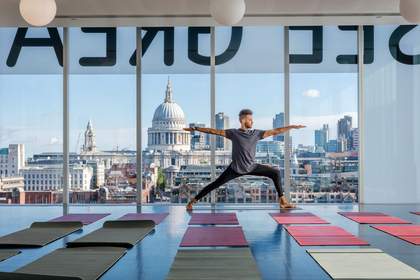 A man is in a yoga pose in a large room with a wall of window.s and a view of St Paul's Cathedral. There are yoga mats on the floor