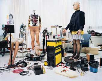 Three robot mannequins stand and sit in a white room with lots of wires on the floor